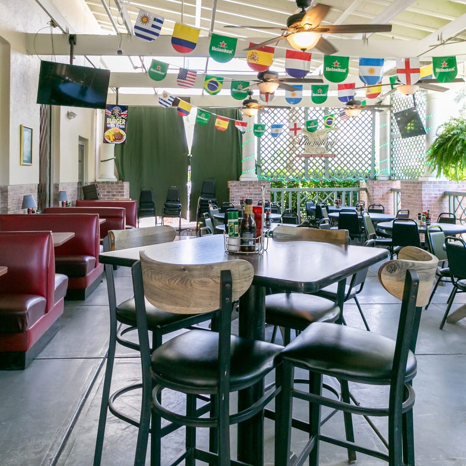 Bright restaurant patio dining area with tables, red booths, ceiling fans, televisions, and international flags hanging overhead.