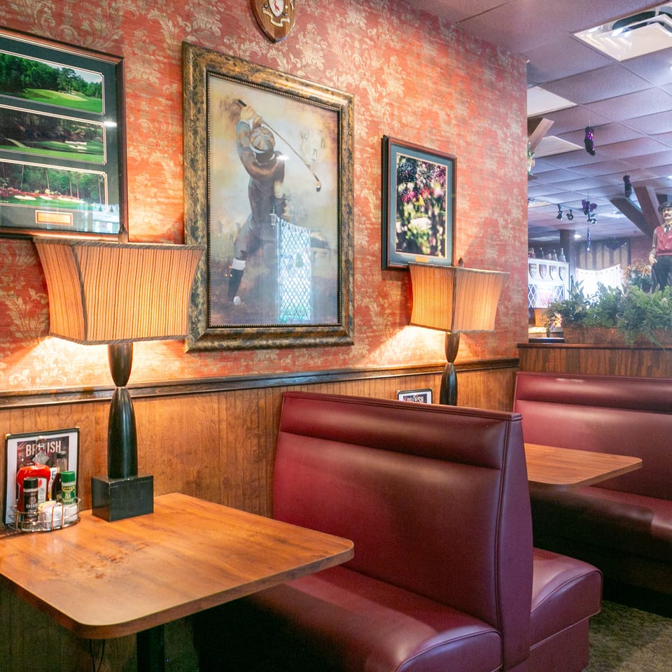 Restaurant dining area with burgundy booths, wooden tables, patterned wallpaper, framed artwork, lamps, and decorative golfer statues.
