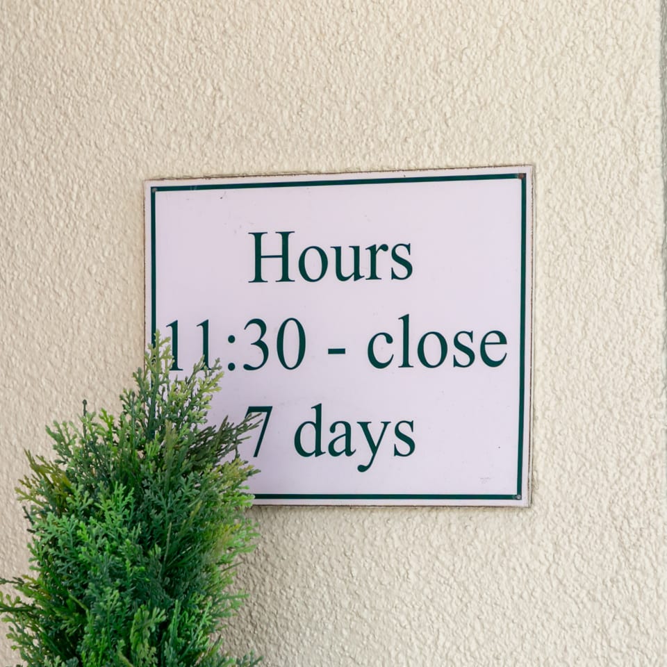 Restaurant hours sign reading “11:30 - close” seven days, mounted on a textured wall beside a potted plant.