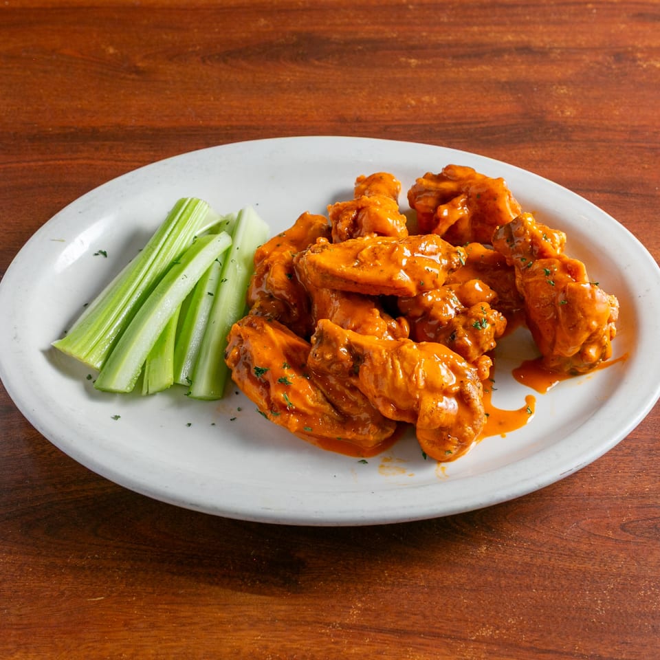 Buffalo chicken wings coated in orange sauce with celery sticks on a white plate atop a wooden table.