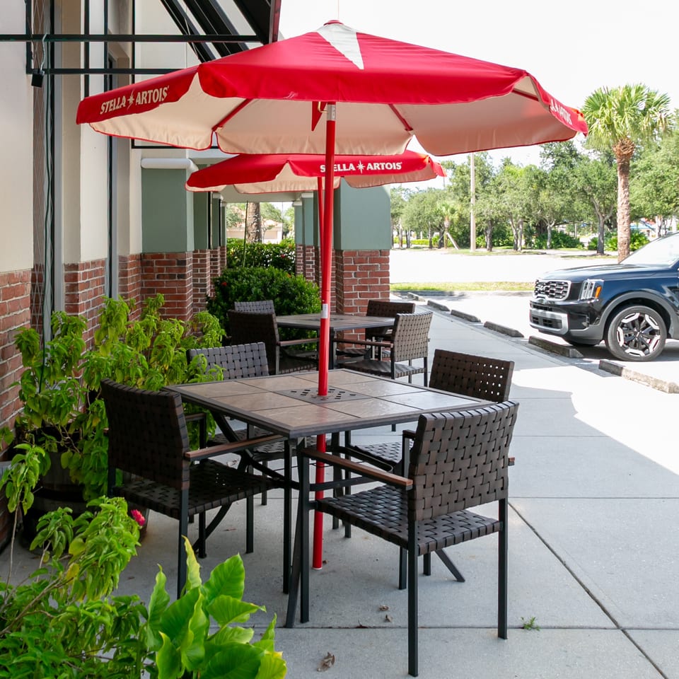 Outdoor restaurant patio with tables, woven chairs, red Stella Artois umbrellas, greenery, and adjacent parking area.