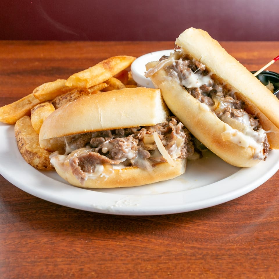 Two cheesesteak sandwiches with sautéed beef and onions served alongside golden fries on a white plate.