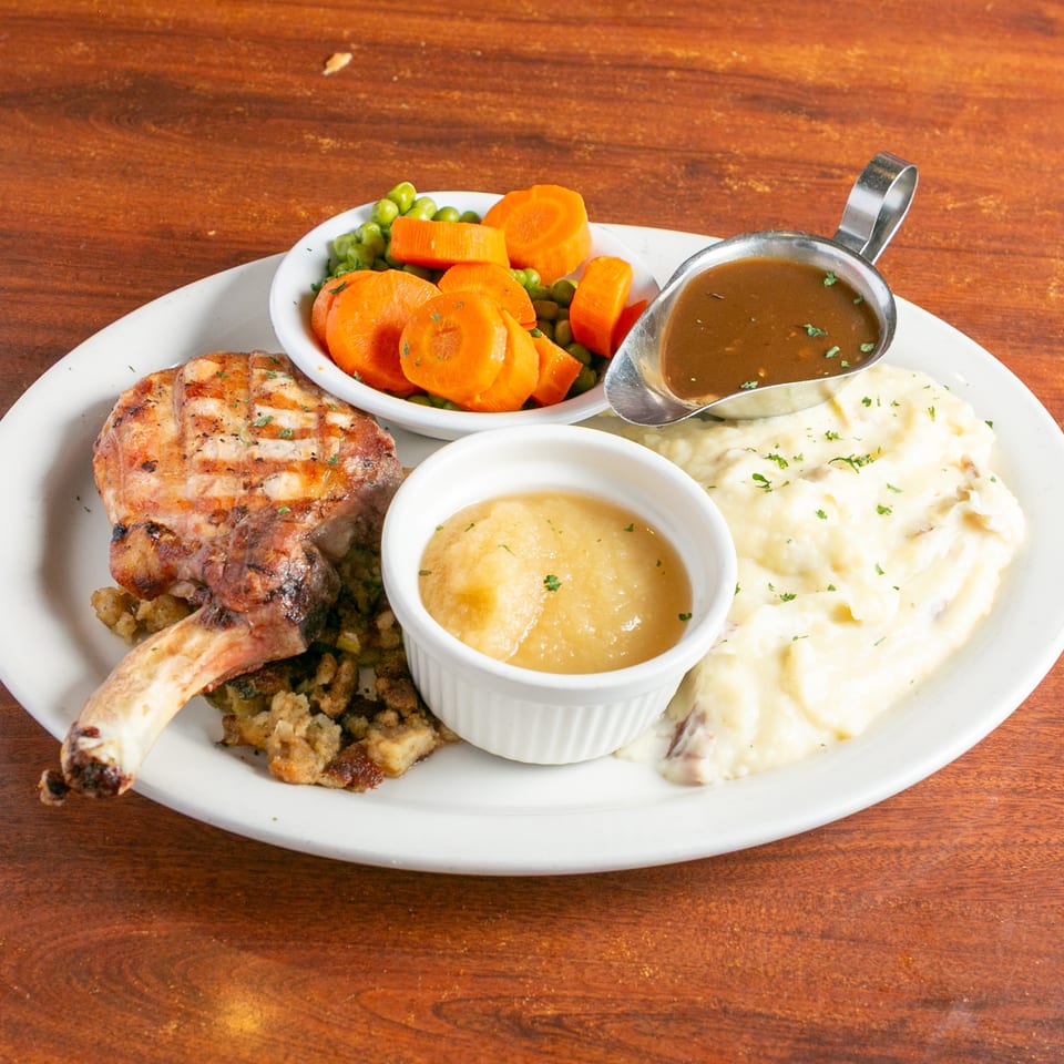 Plated comfort-food dinner featuring grilled meat, stuffing, mashed potatoes, gravy, applesauce, and peas with carrots.
