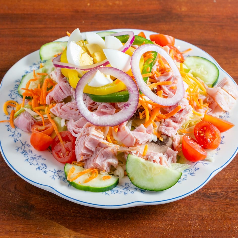 Colorful ham and vegetable salad with egg, onions, carrots, tomatoes, cucumbers, and peppers on a patterned plate.