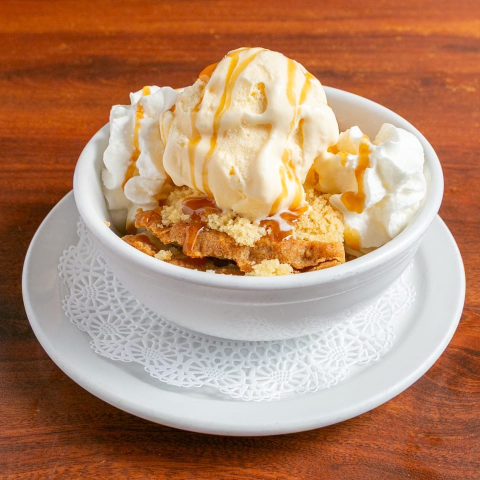 Bowl of crispy dessert topped with vanilla ice cream, whipped cream, and caramel sauce on a white plate.